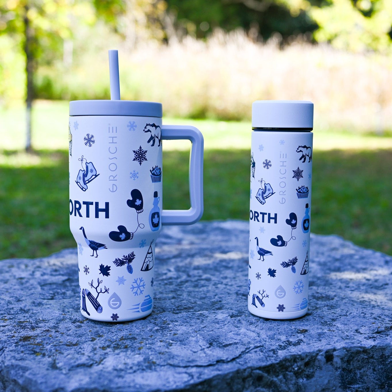 Two insulated travel mugs with winter-themed designs on a stone surface outdoors.