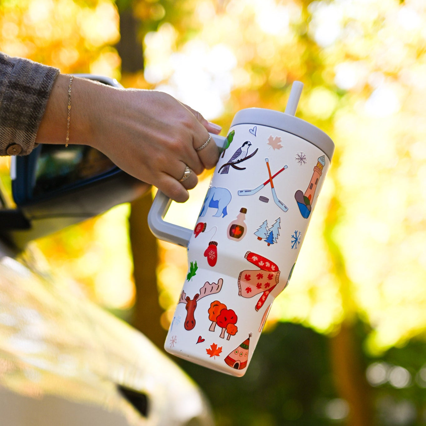 Person holding a colorful tumbler with Canadian-themed illustrations out of a car window in an outdoor setting.
