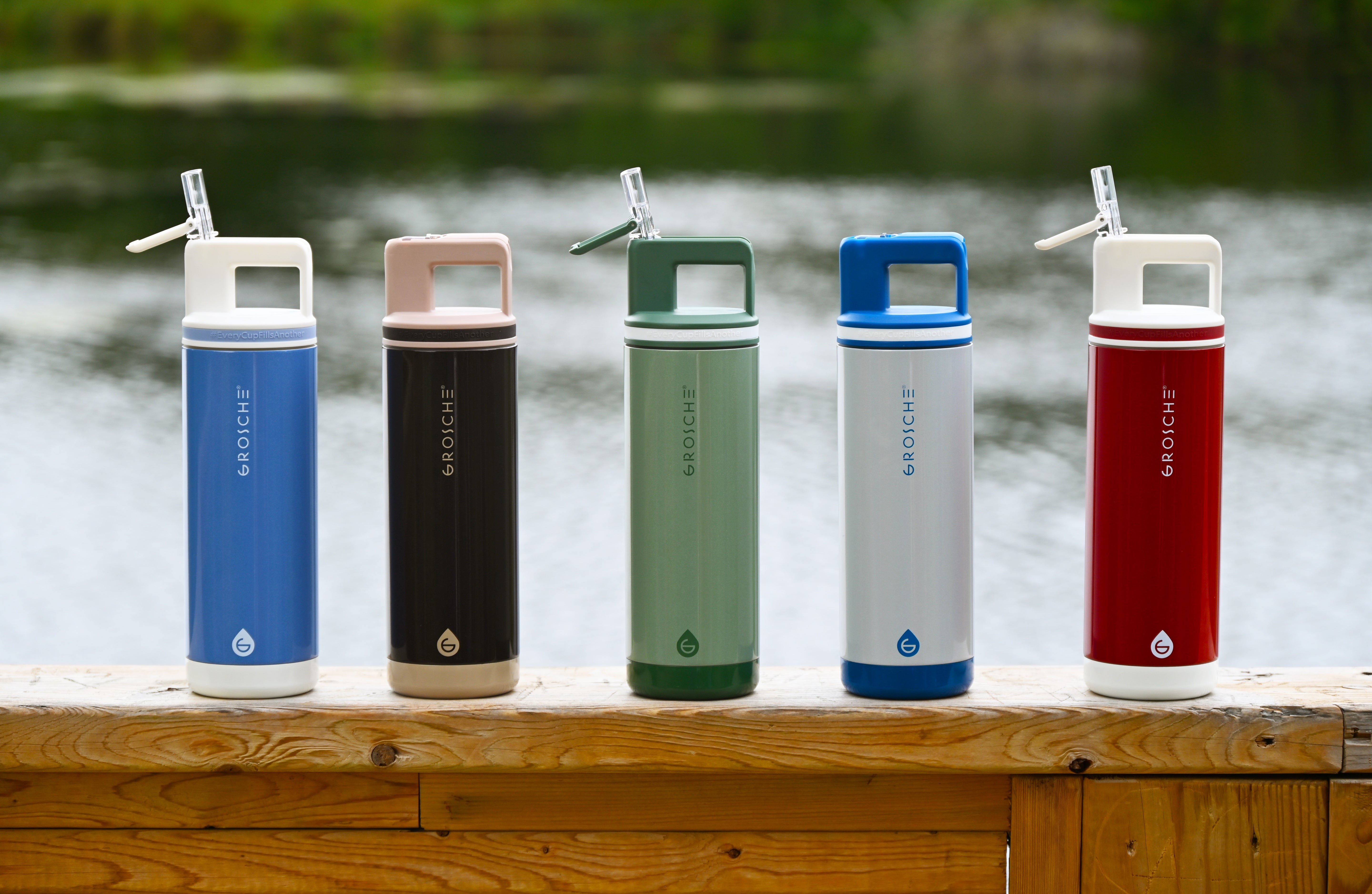 Five colorful water bottles with straws on a wooden surface by a body of water.