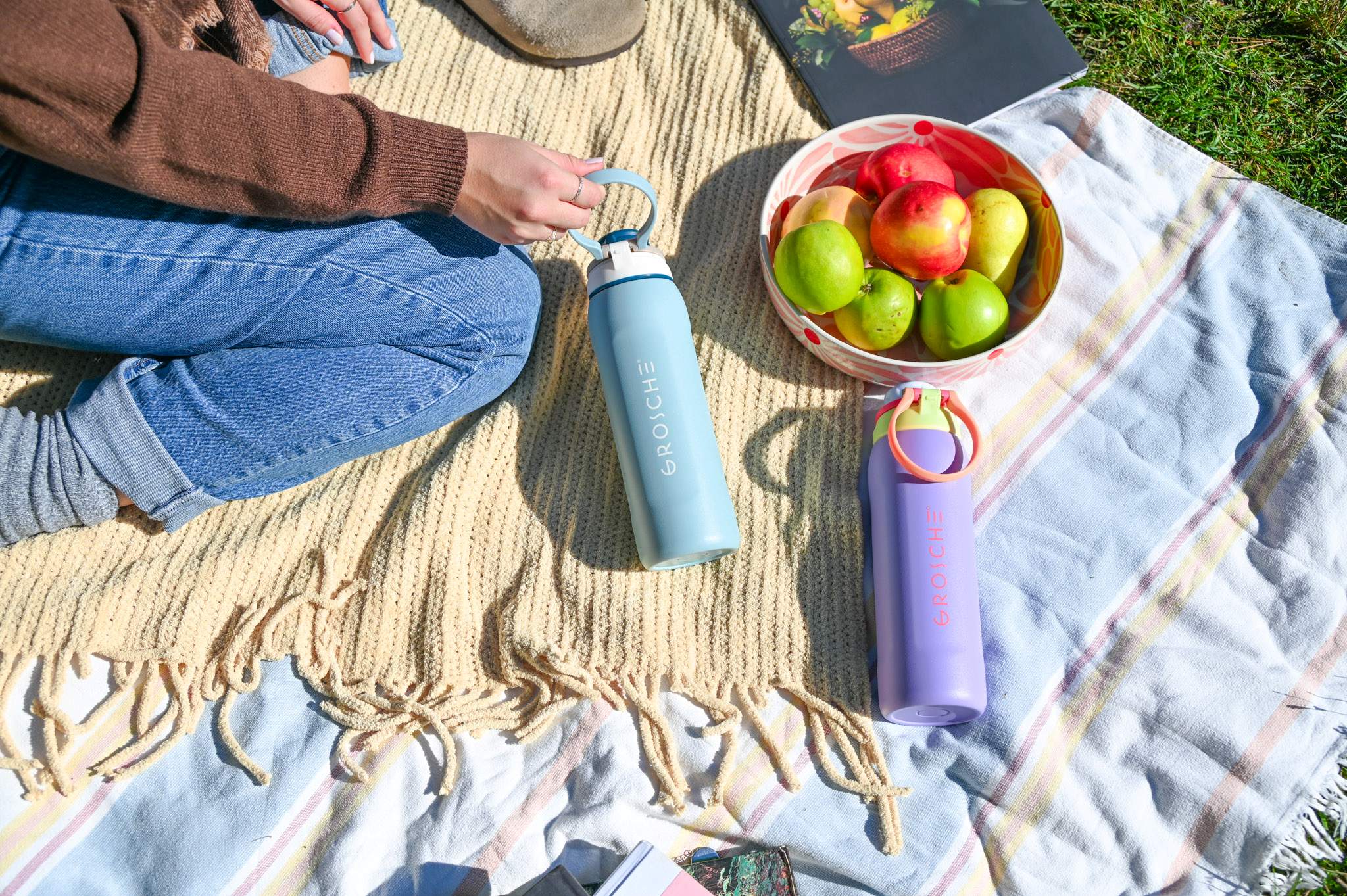 GROSCHE Kamloops Person sitting on a blanket with a blue water bottle and a purple water bottle with a bowl of fruit.