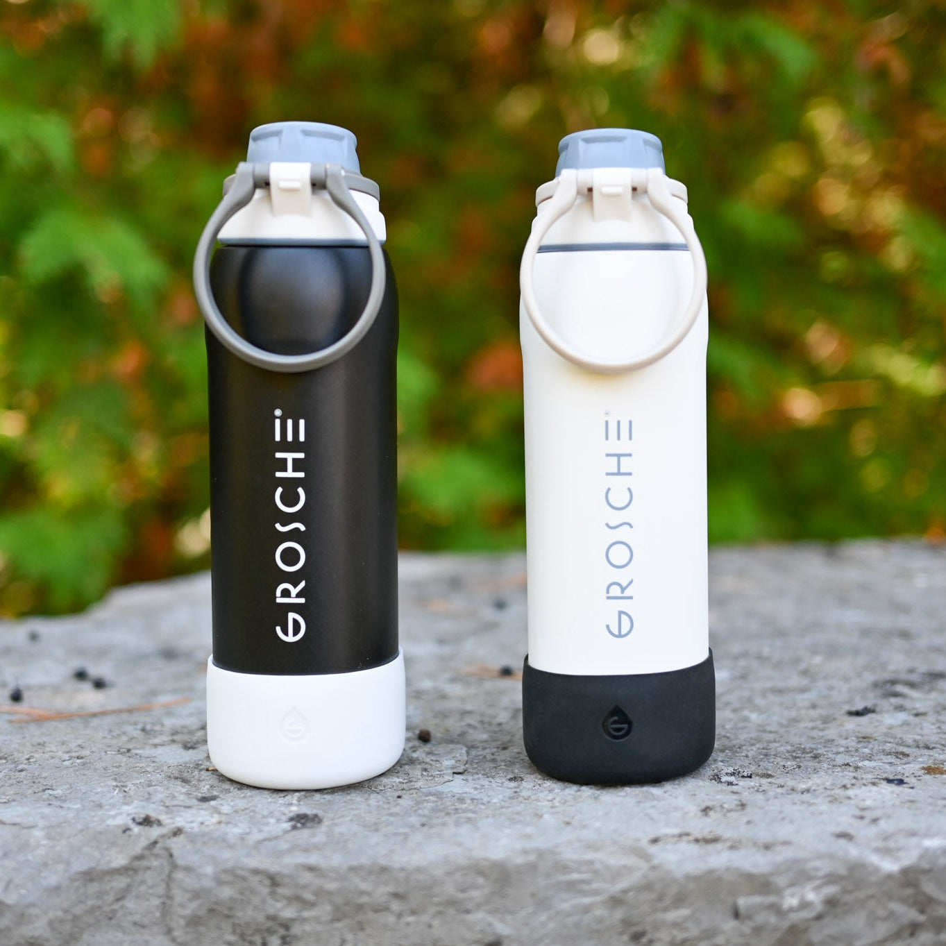 Two GROSCHE bottles, one black and one white, on a stone surface with a blurred green background.
