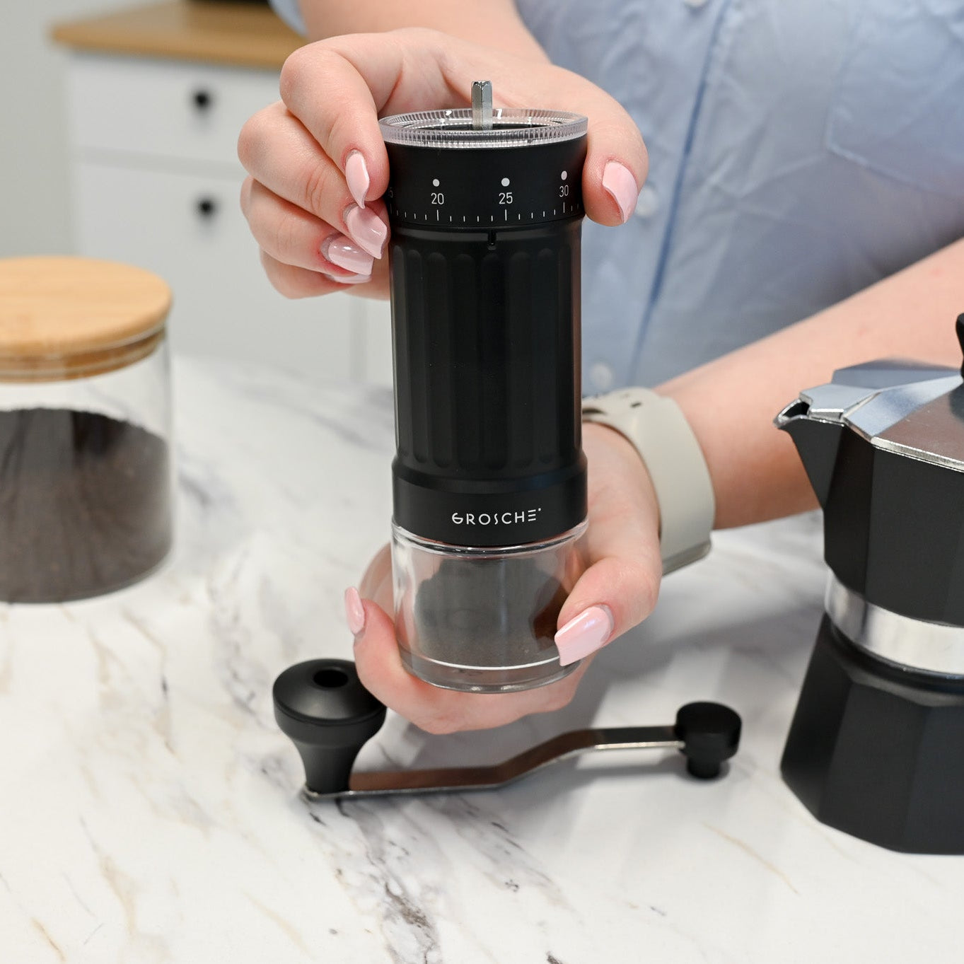 Person holding a black coffee grinder with a GROSCHE Moka pot on a marble surface.