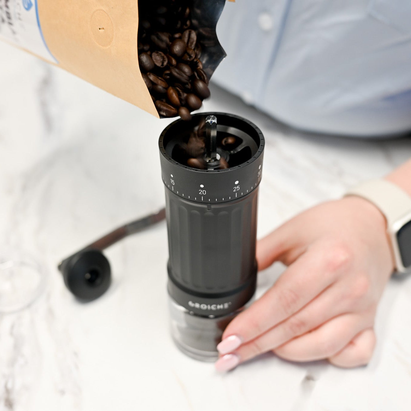 Person pouring coffee beans into a manual coffee grinder on a white surface.