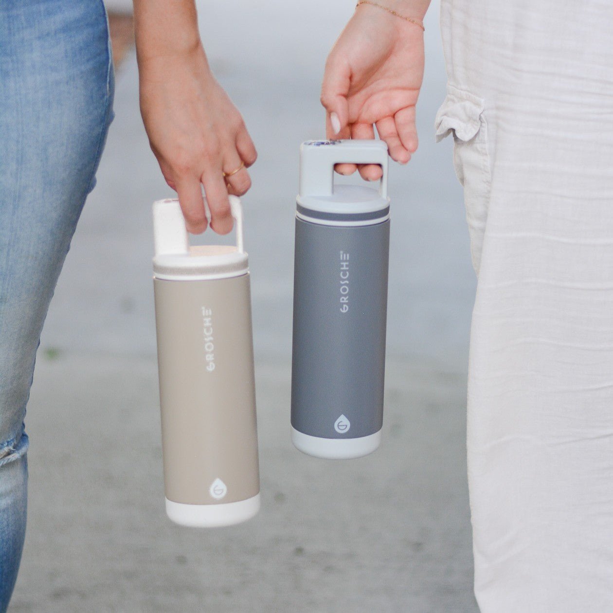 Two people each holding a GROSCHE Alpine water bottle. One is beige with a white lid and the other is blue with a light blue lid. 