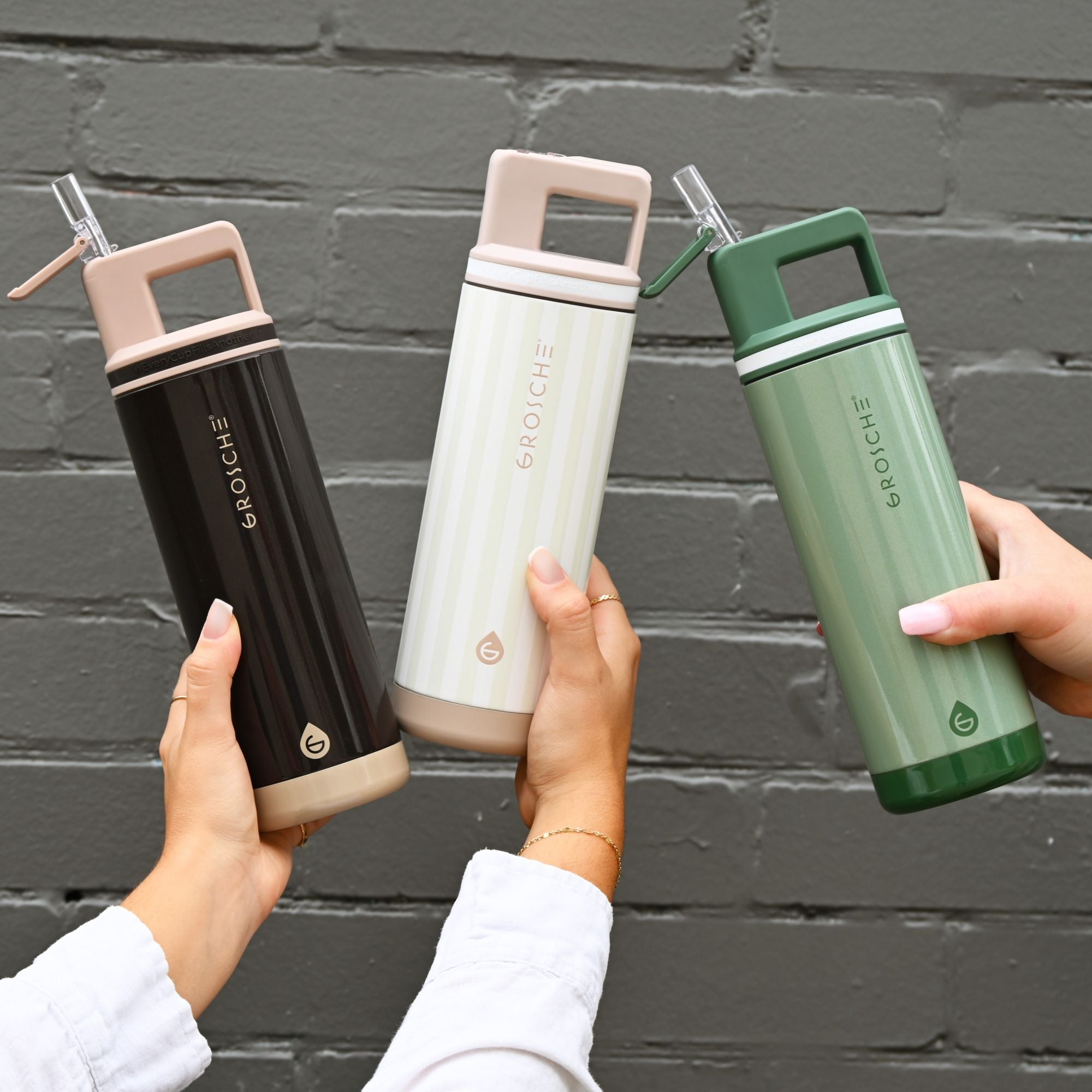 Three hands holding black, white, and green water bottles against a gray brick wall.
