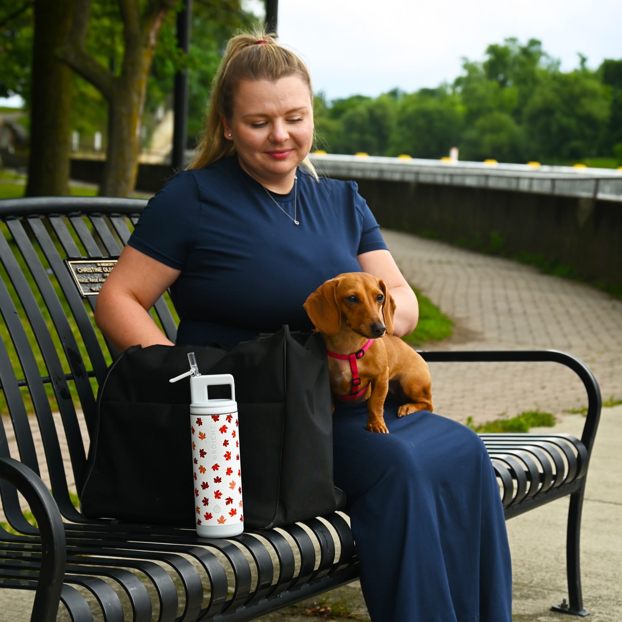 Woman sitting on a bench with a small dog, holding a water bottle with a park in the background