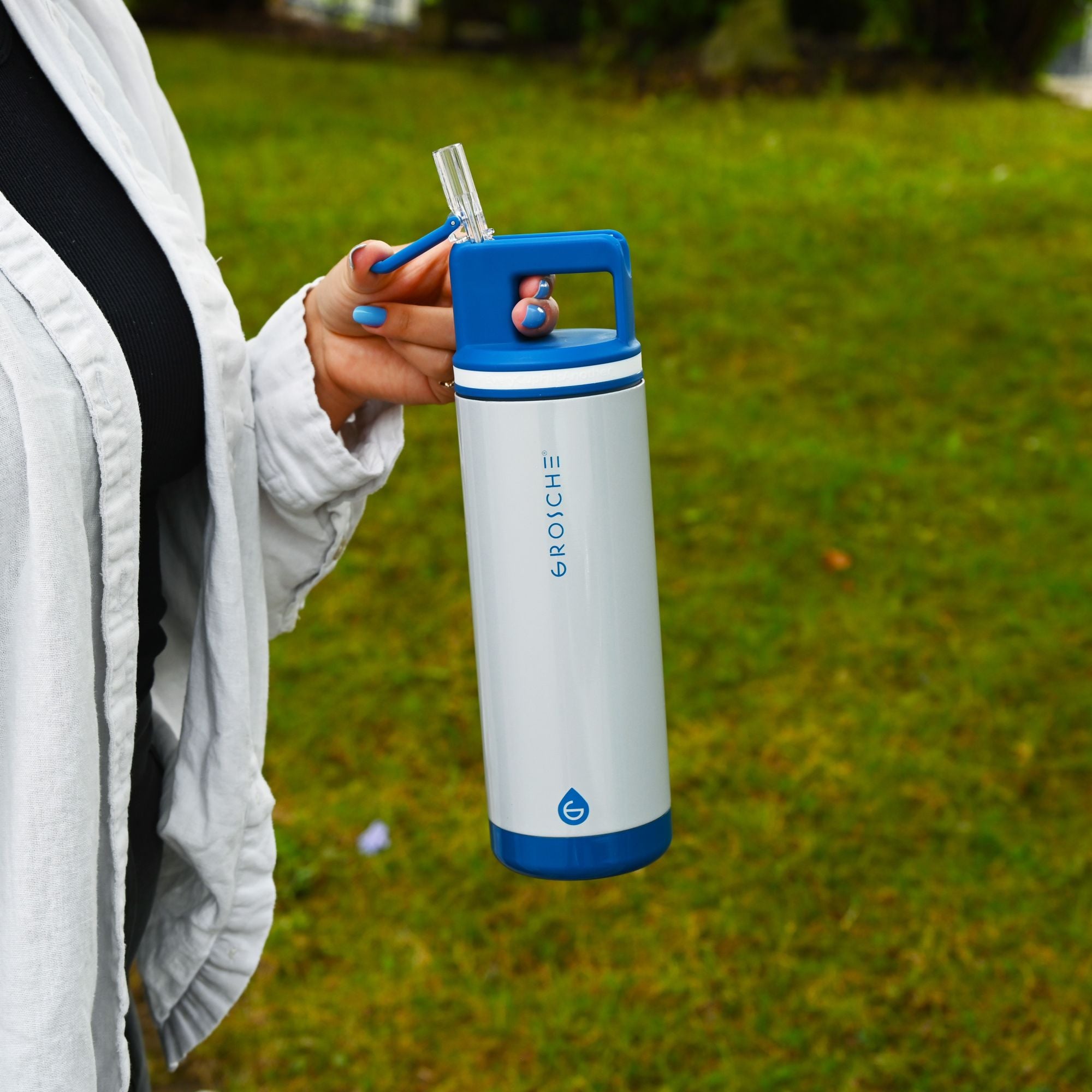 Person holding a white and blue GROSCHE Alpine water bottle with a straw lid outdoors on grass