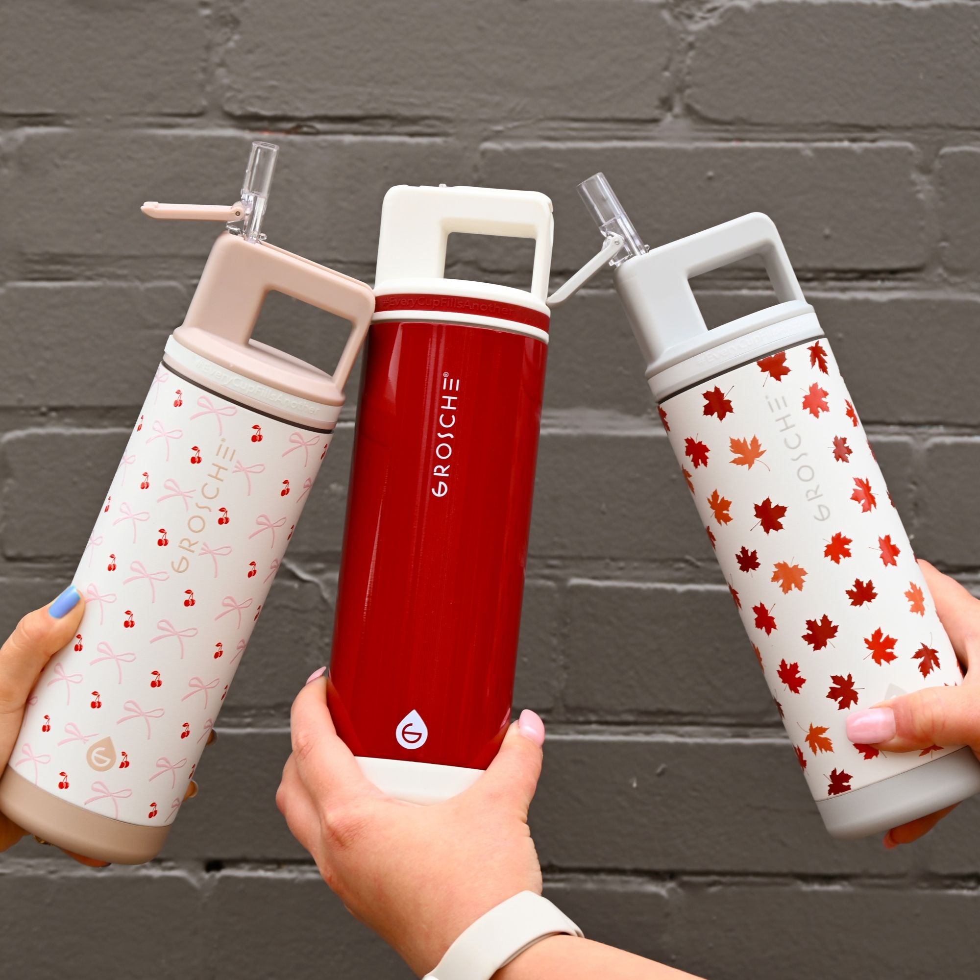 Three GROSCHE water bottles with different designs held against a gray brick wall.