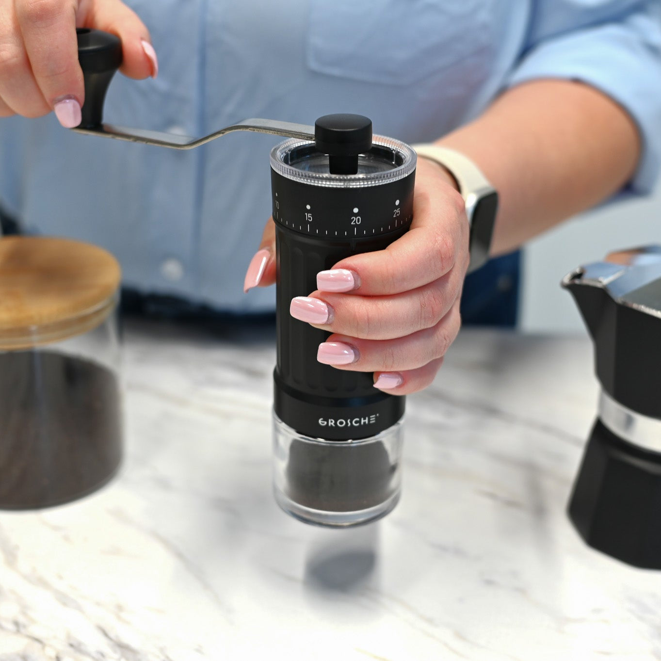 Person using a manual coffee grinder with a Grosche Moka pot in the background