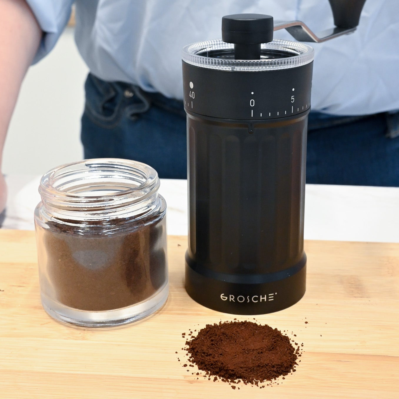 Black manual coffee grinder with ground coffee on a wooden surface