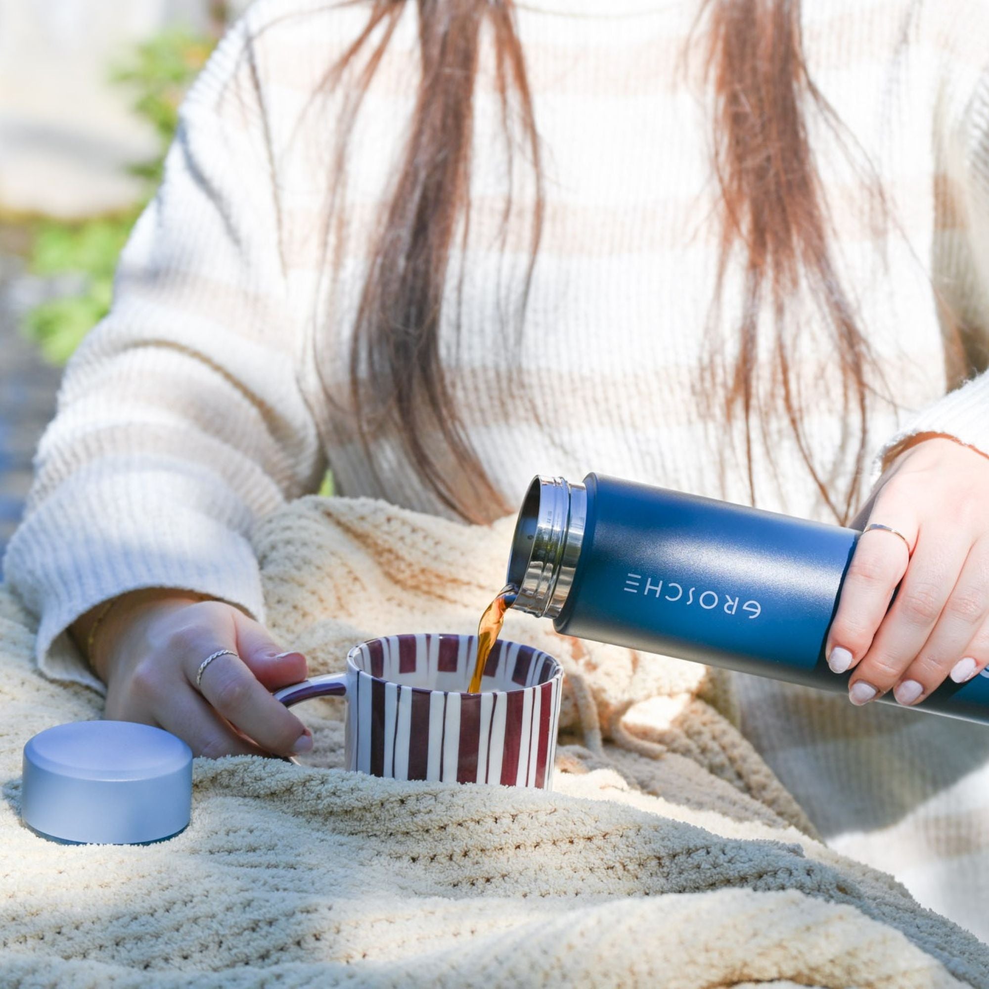 Person pouring coffee from a Grosche thermos into a striped mug outdoors.