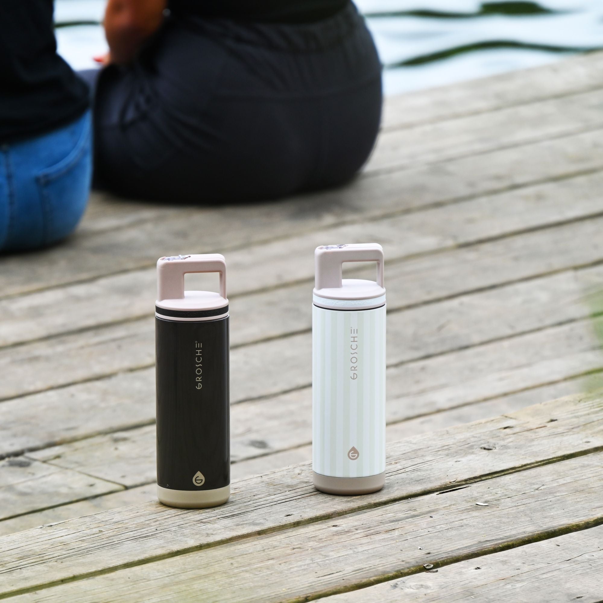 Two GROSCHE  Alpine water bottles on a wooden surface with two people blurred in the background. 